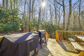 Eine Holzterrasse mit Grill und Picknicktisch, umgeben von Bäumen und Grünpflanzen.