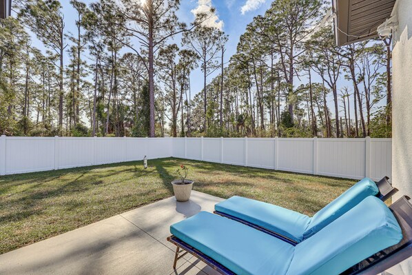 An outdoor patio with two blue lounge chairs, a concrete floor, a white fence, and a backdrop of tall pine trees. - Stone's Throw to All Major Attractions: Family Gem (North Port)