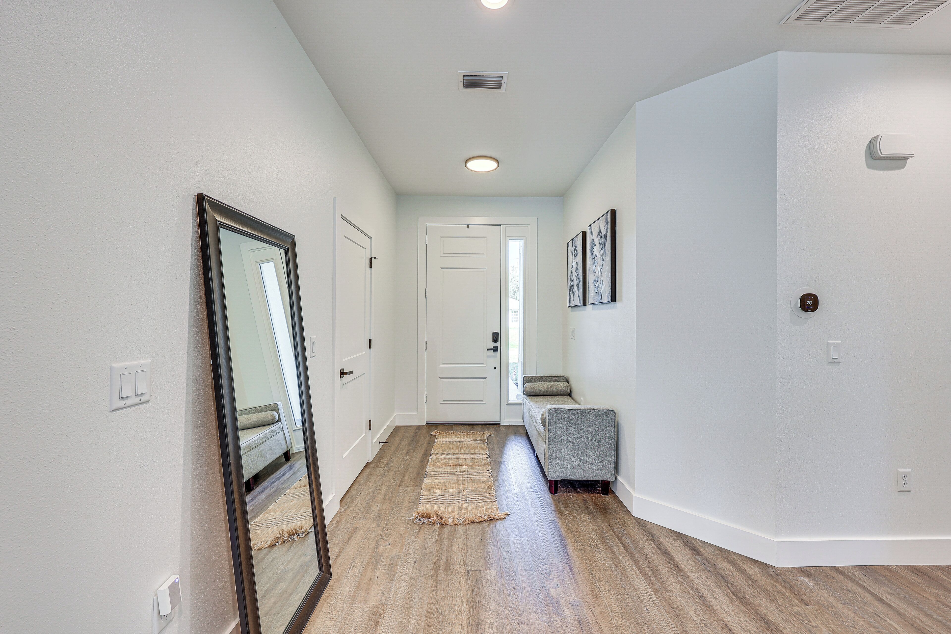A hallway with hardwood floors, white walls, a mirror, a door, and a gray bench.