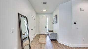 A hallway with hardwood floors, white walls, a mirror, a door, and a gray bench.