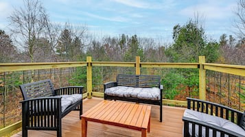 Una terraza de madera con dos bancos negros y una mesa, rodeada por una barandilla amarilla y una cerca de alambre.