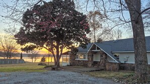 A stone house with a covered porch, a gravel driveway, and a view of a lake at sunset. - Deck & Yard: Wolf Creek Waterfront Home in Grove! (Grove)