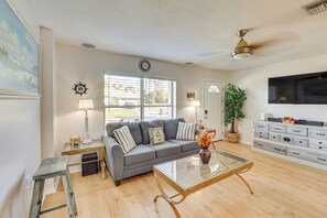 A living room with a gray sofa, a glass coffee table, a side table with a lamp, a wall clock, and a television on the wall. - Summer-ready North Port Home w/ Private Pool! (North Port)