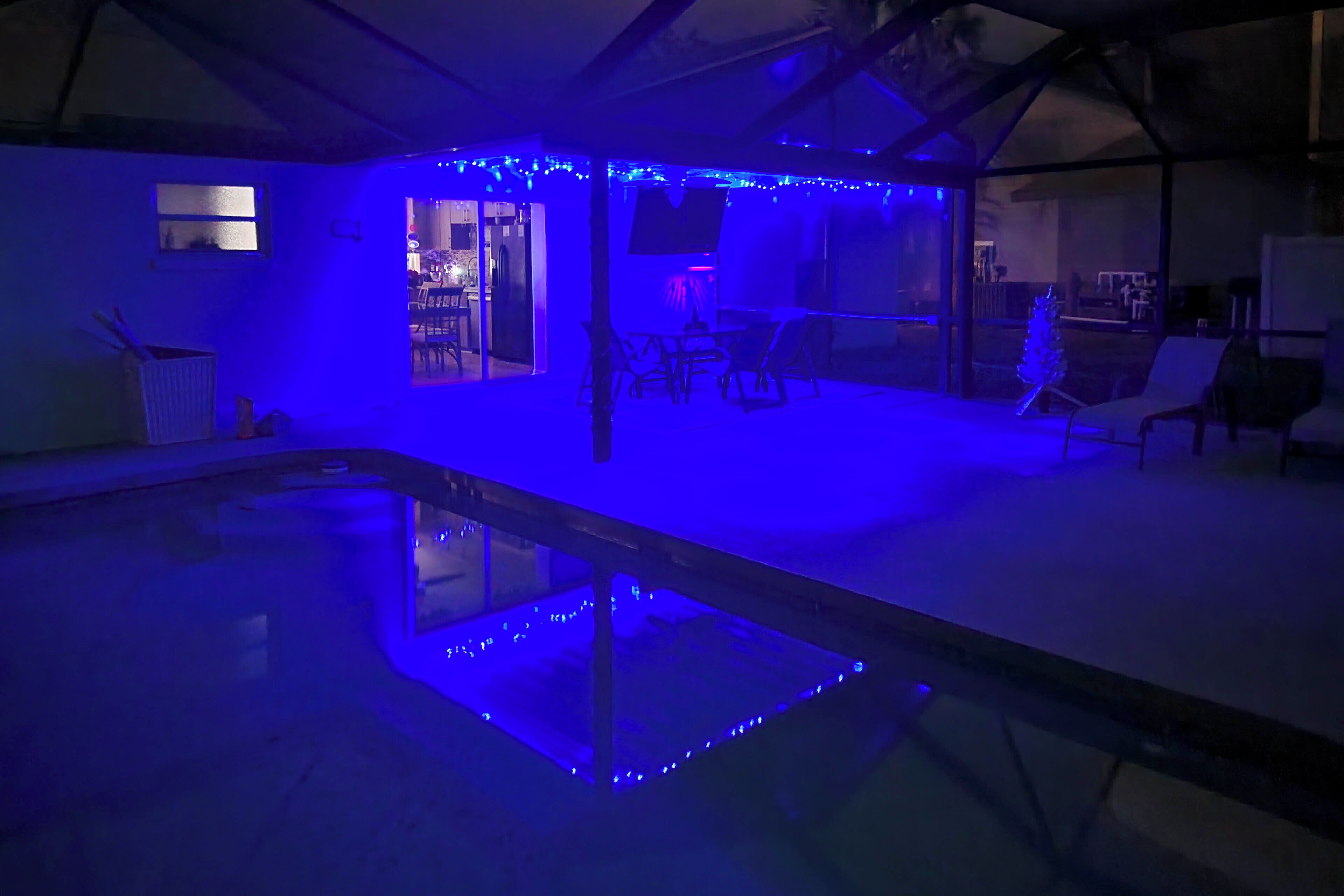 Un espace piscine avec un éclairage d’ambiance bleuté, un coin salon avec des chaises, et une entrée visible menant à une autre pièce.
