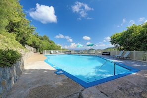 Una piscina rectangular con borde azul, rodeada por un muro de piedra y una terraza de madera. Se ven dos sombrillas y sillones de descanso al fondo.