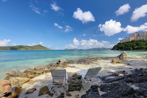 Una playa con dos sillas reclinables blancas orientadas al mar, agua turquesa cristalina y un edificio en una colina al fondo.