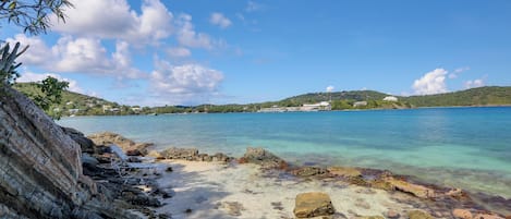 A beach with clear turquoise water, rocky shoreline, and a distant view of a hill with buildings.
