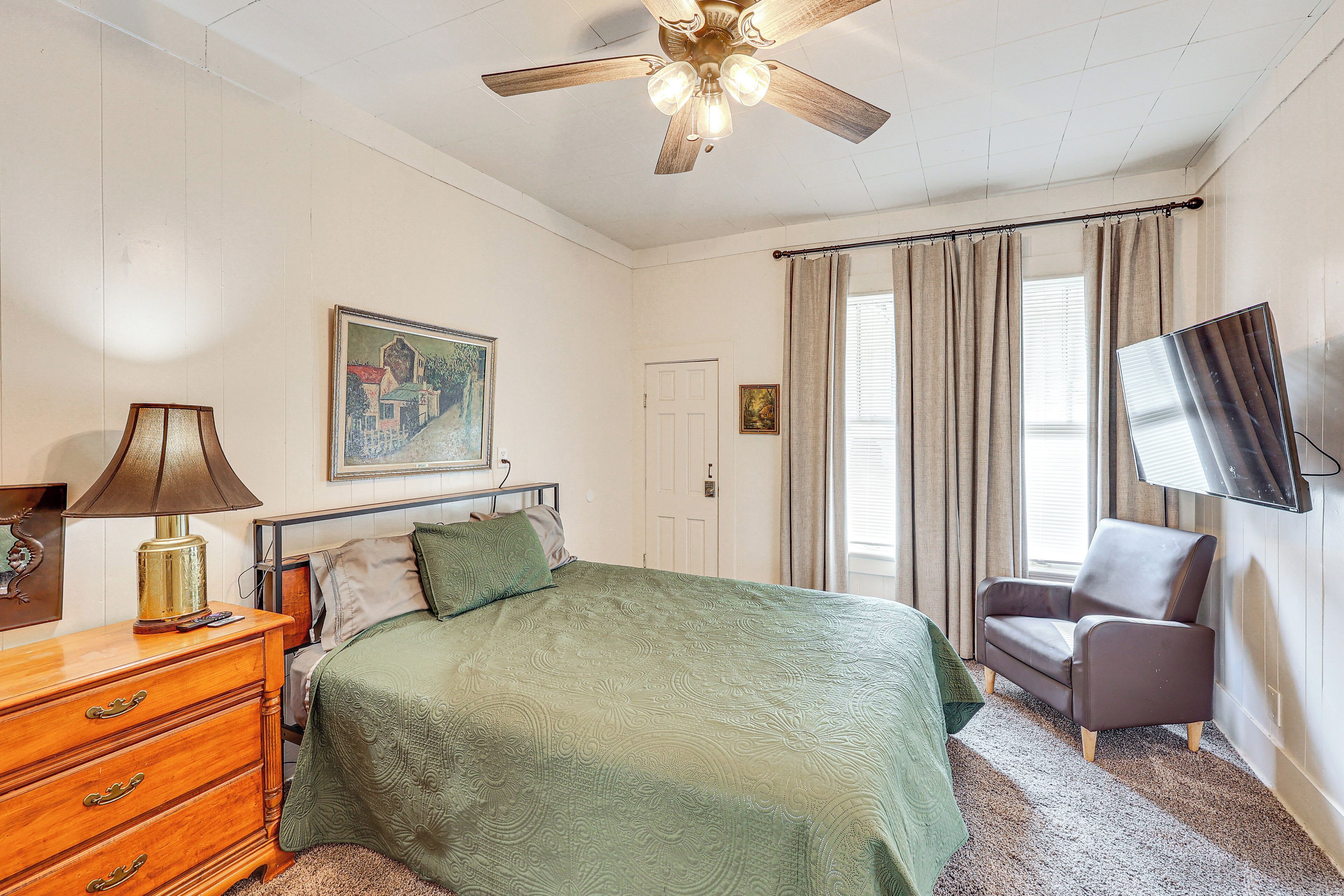 A bedroom with a bed, a dresser, a chair, a ceiling fan, a TV mounted on the wall, and a window with curtains.