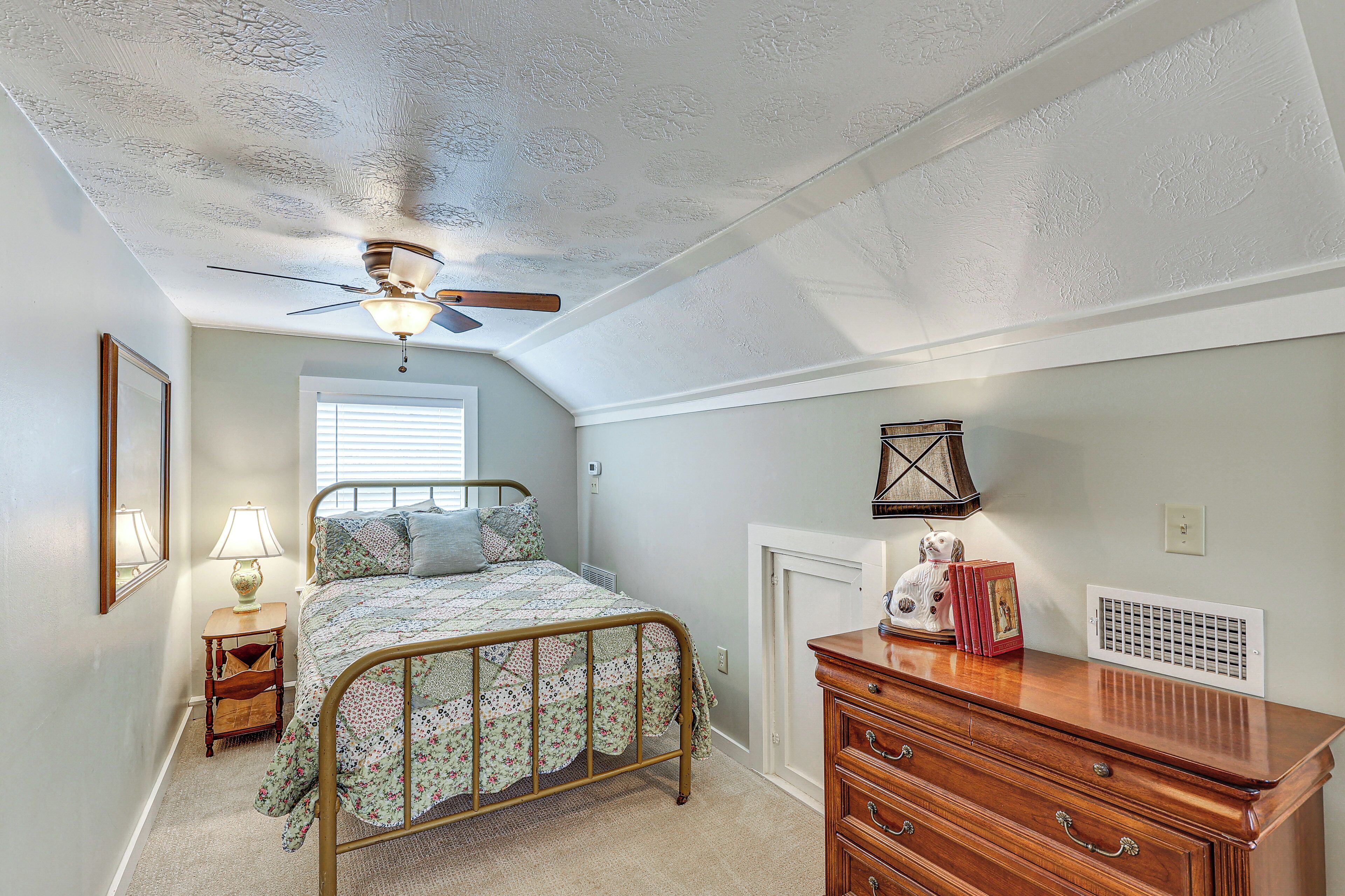 A bedroom with a bed, dresser, nightstand, lamp, ceiling fan, and a radiator.