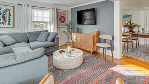 A modern living room with a grey sectional sofa, a round coffee table, and a large flat-screen TV mounted on the wall. - Escape to Cape Ann Steps to Beach & Pier! (Gloucester)