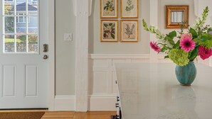 A room with a white door, a white wall with framed botanical illustrations, and a vase with flowers on a marble table. - Escape to Cape Ann Steps to Beach & Pier! (Gloucester)