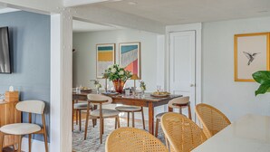 A dining area with a wooden table, wicker chairs, and a vase with flowers. There are framed artworks on the wall and a television mounted on the wall. - Escape to Cape Ann Steps to Beach & Pier! (Gloucester)