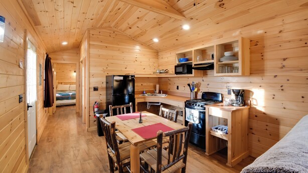 A wooden cabin interior with a dining area, kitchen, and a bed. - Shared Dock Access at Lakeside Sodus Bay Cabin! (North Rose)
