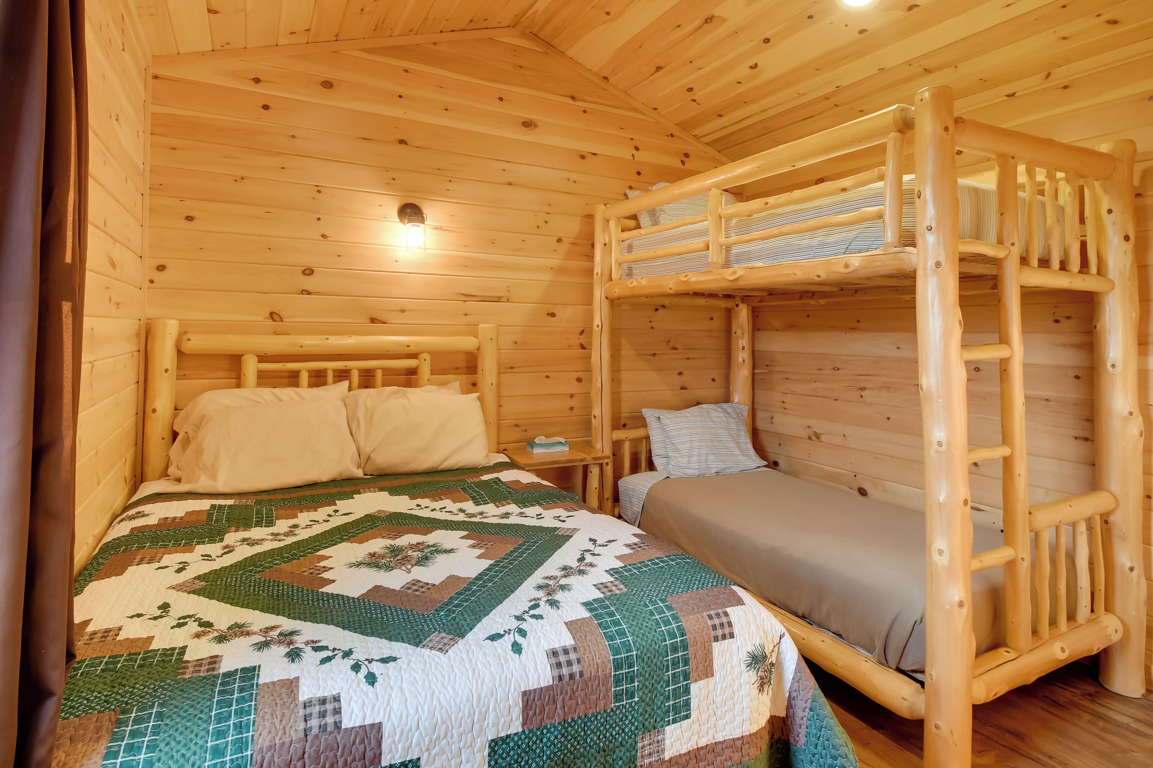 A wooden cabin room with bunk beds, a quilted bedspread, and a small bedside table with a lamp.