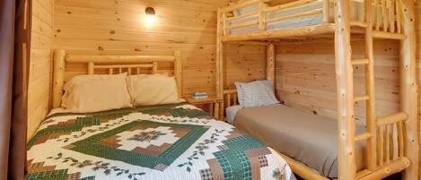 A wooden cabin room with bunk beds, a quilted bedspread, and a small bedside table with a lamp.