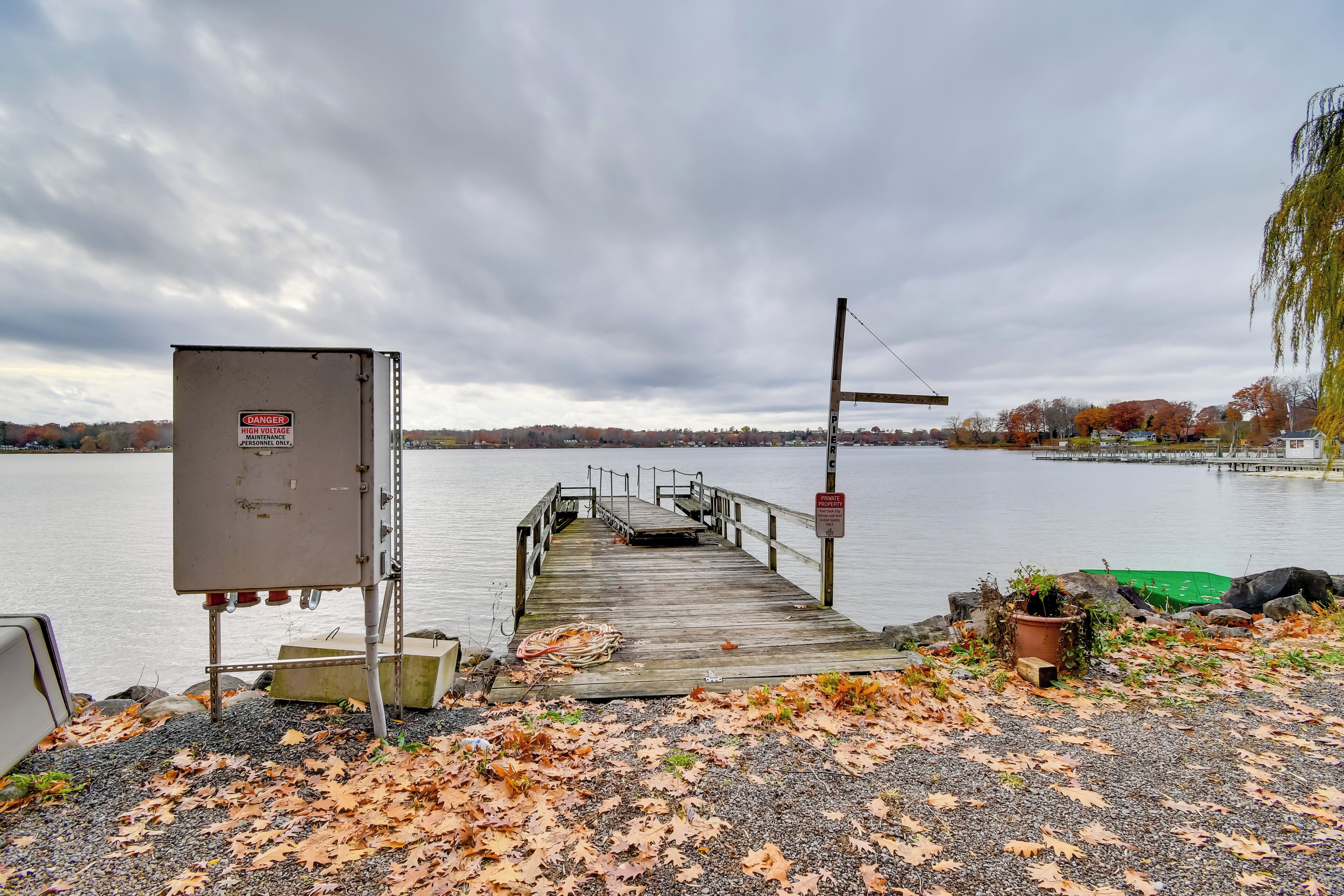 A wooden dock extending into a lake with a 'Danger High Voltage' sign, a utility box, and a utility pole with a sign.