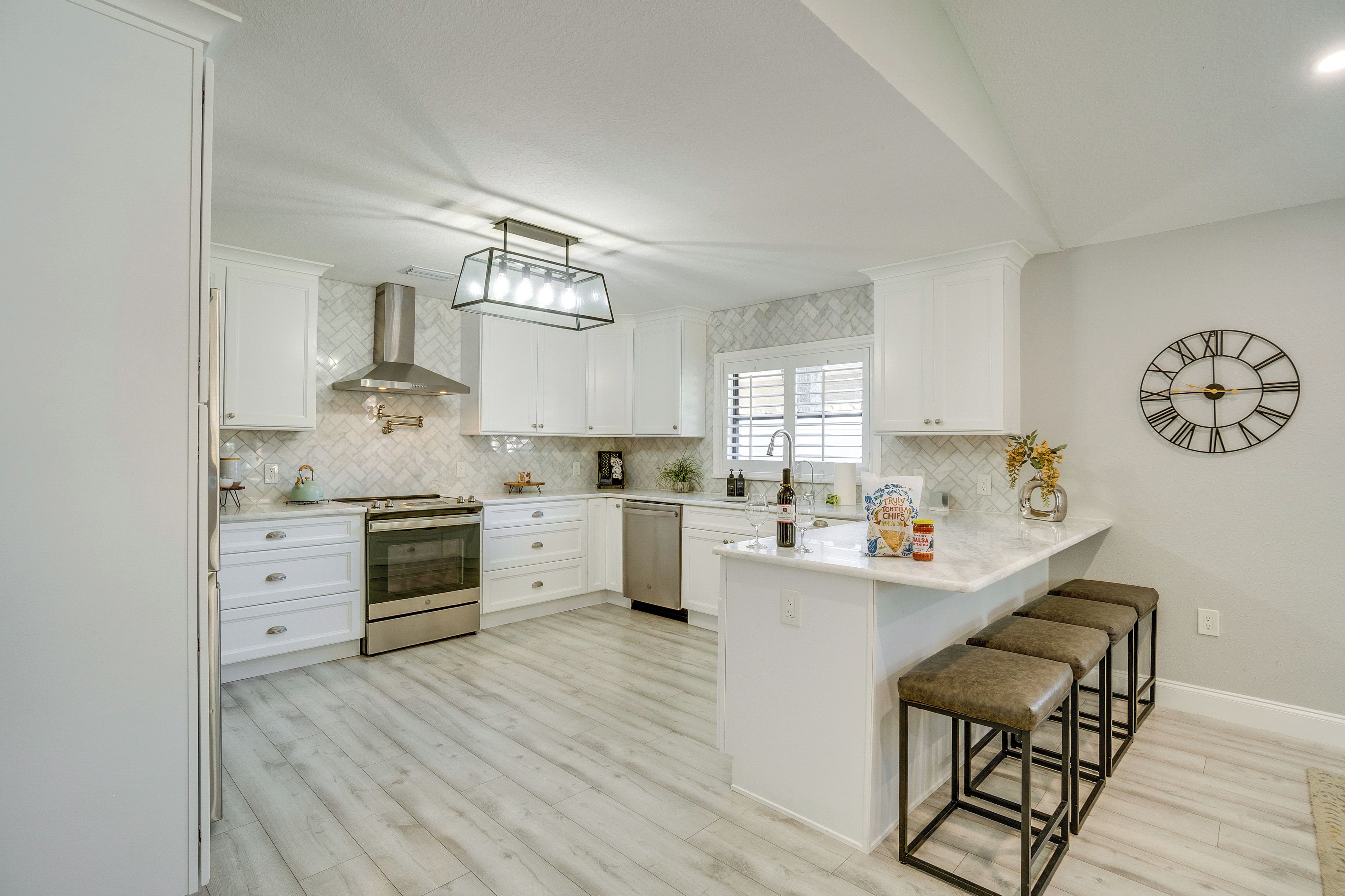 A modern kitchen with white cabinetry, a central island with stools, stainless steel appliances, and a decorative clock on the wall.