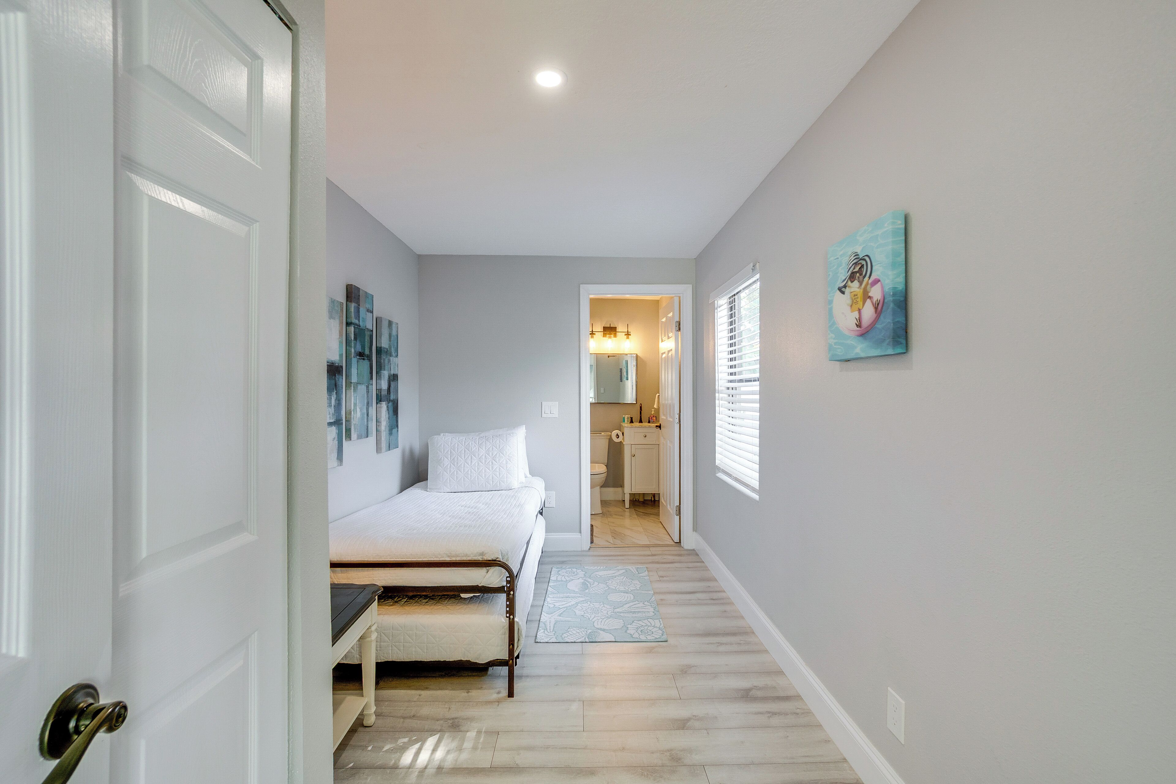Un dormitorio con cama, mesita de noche, persianas en la ventana y una puerta que da a otra habitación.