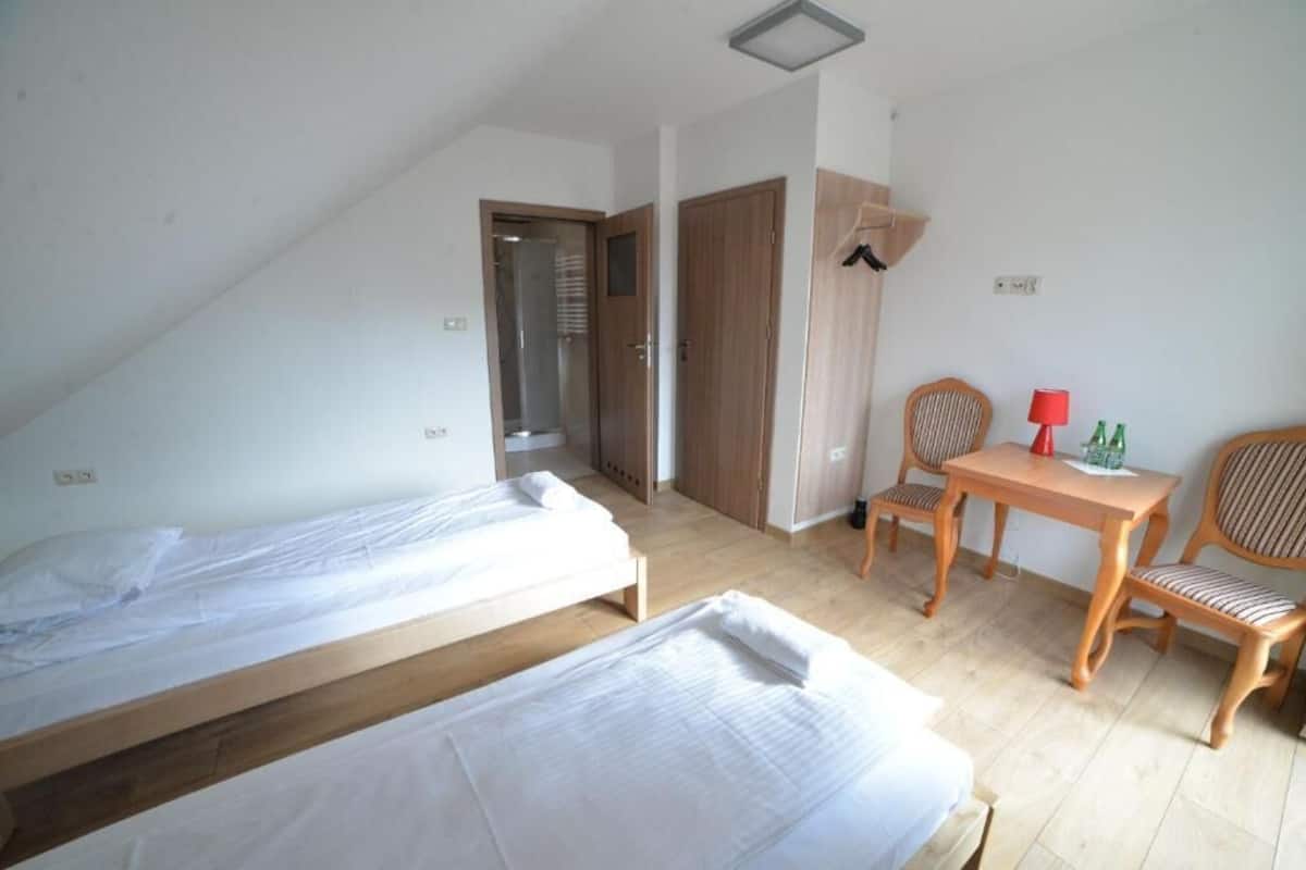 A room with two single beds, a wooden table with chairs, a red lamp, and a small table with bottles.
