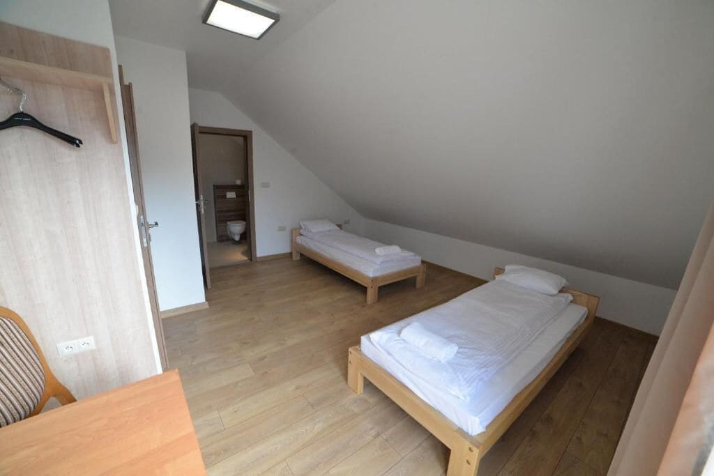 Une petite pièce bien éclairée, avec deux lits en bois, une chaise en bois et une porte donnant sur une salle de bain.