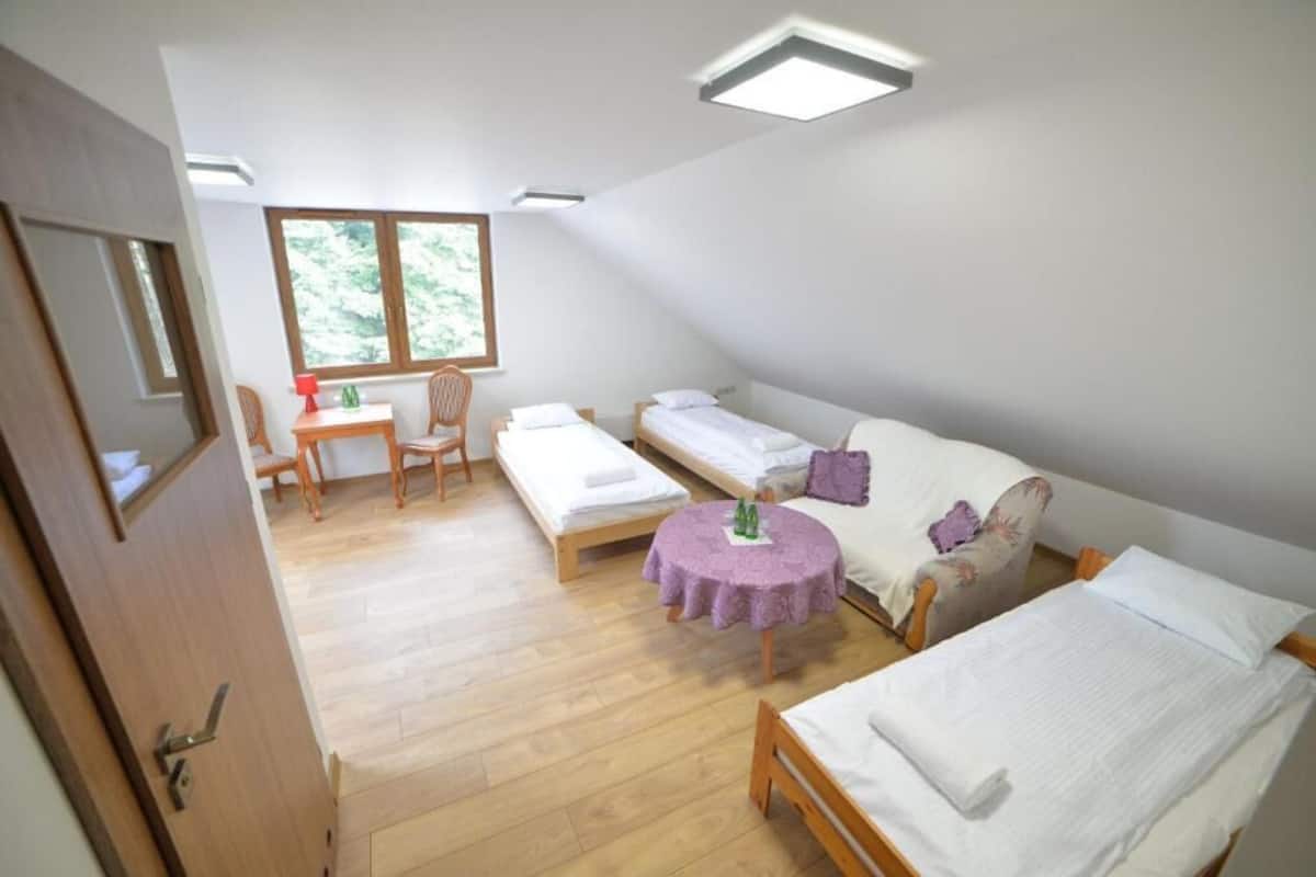 A dormitory room with two single beds, a sofa bed, a small round table with a purple tablecloth, and a window providing natural light.