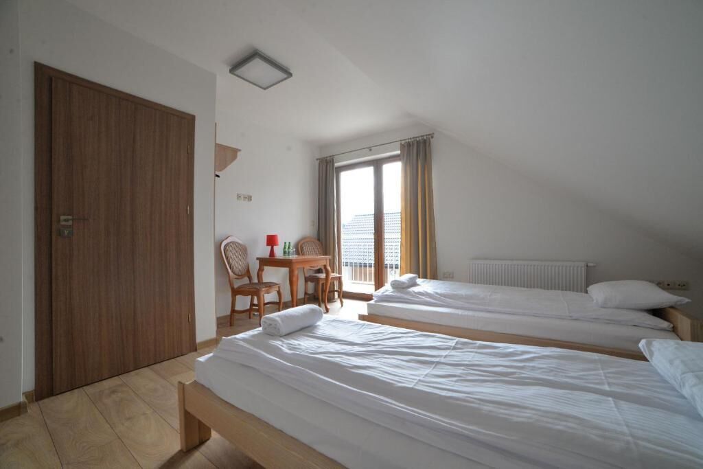 A hotel room with two beds, a wooden door, a small table with chairs, and a window with curtains.