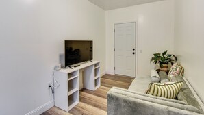 A living room with a flat-screen TV on a white stand, a grey sofa with striped cushions, and a potted plant.