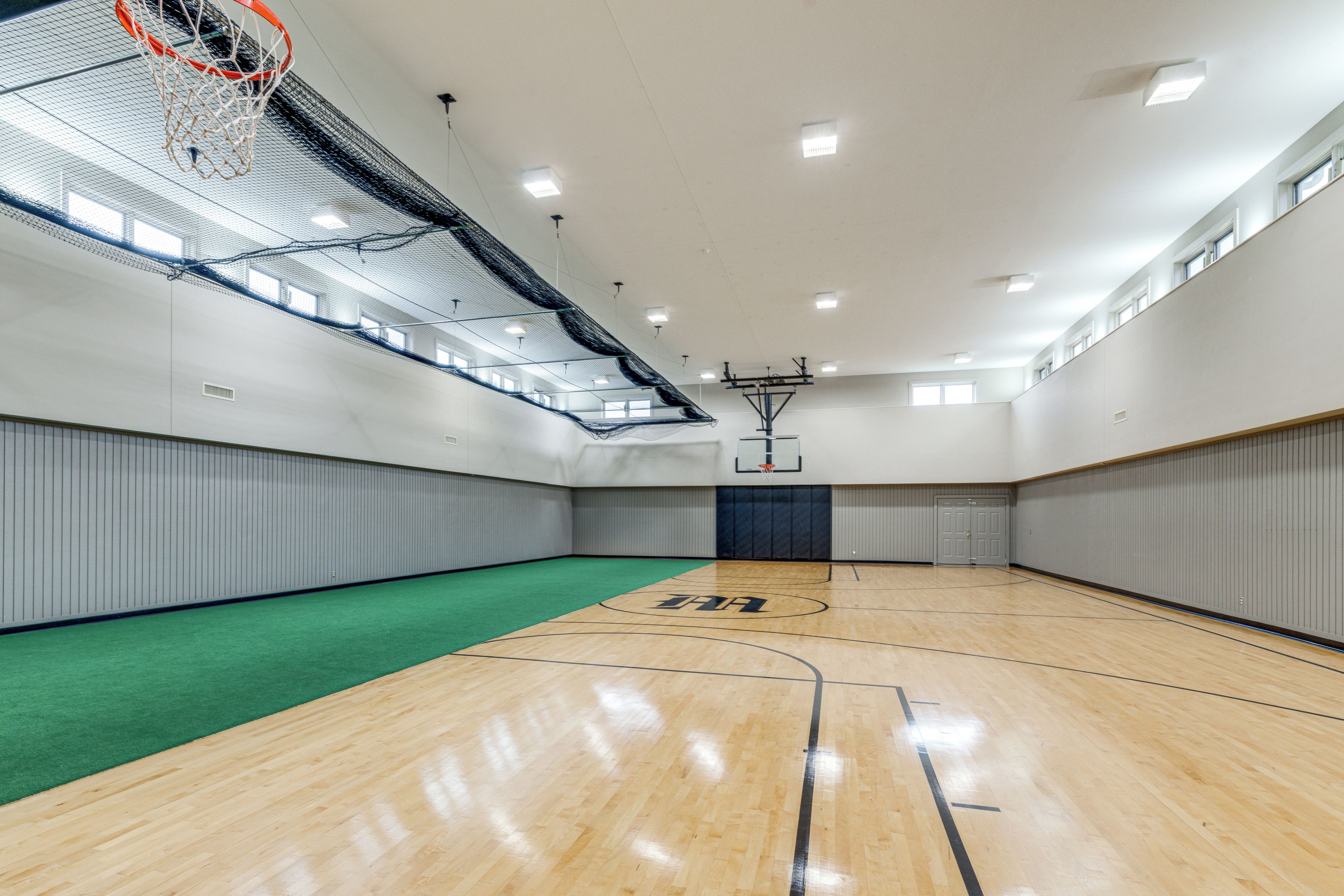 A spacious indoor basketball court with a wooden floor, a basketball hoop, and a green carpeted area.
