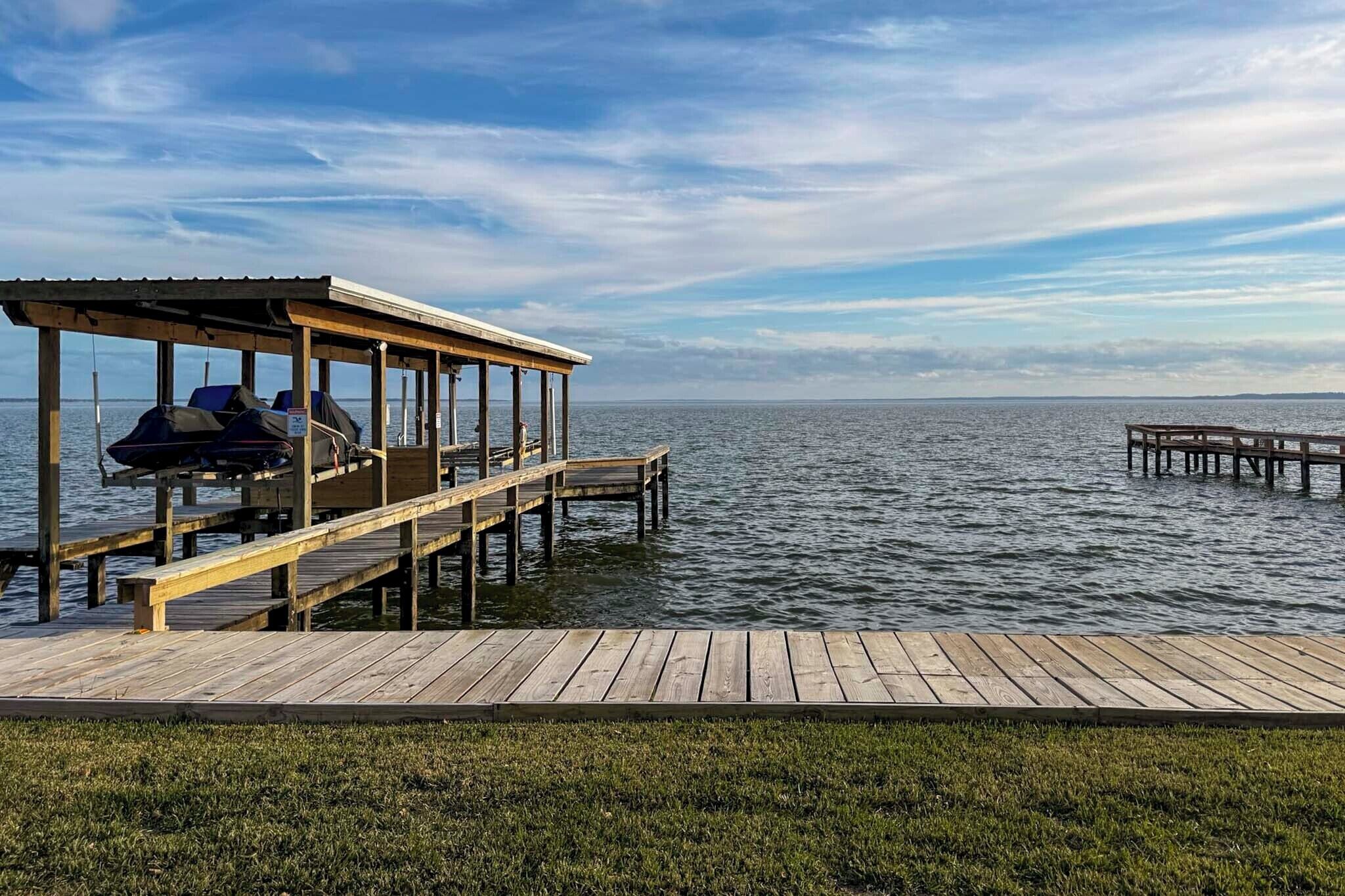 A wooden dock extending into calm water, with a covered seating area and grill on the left.