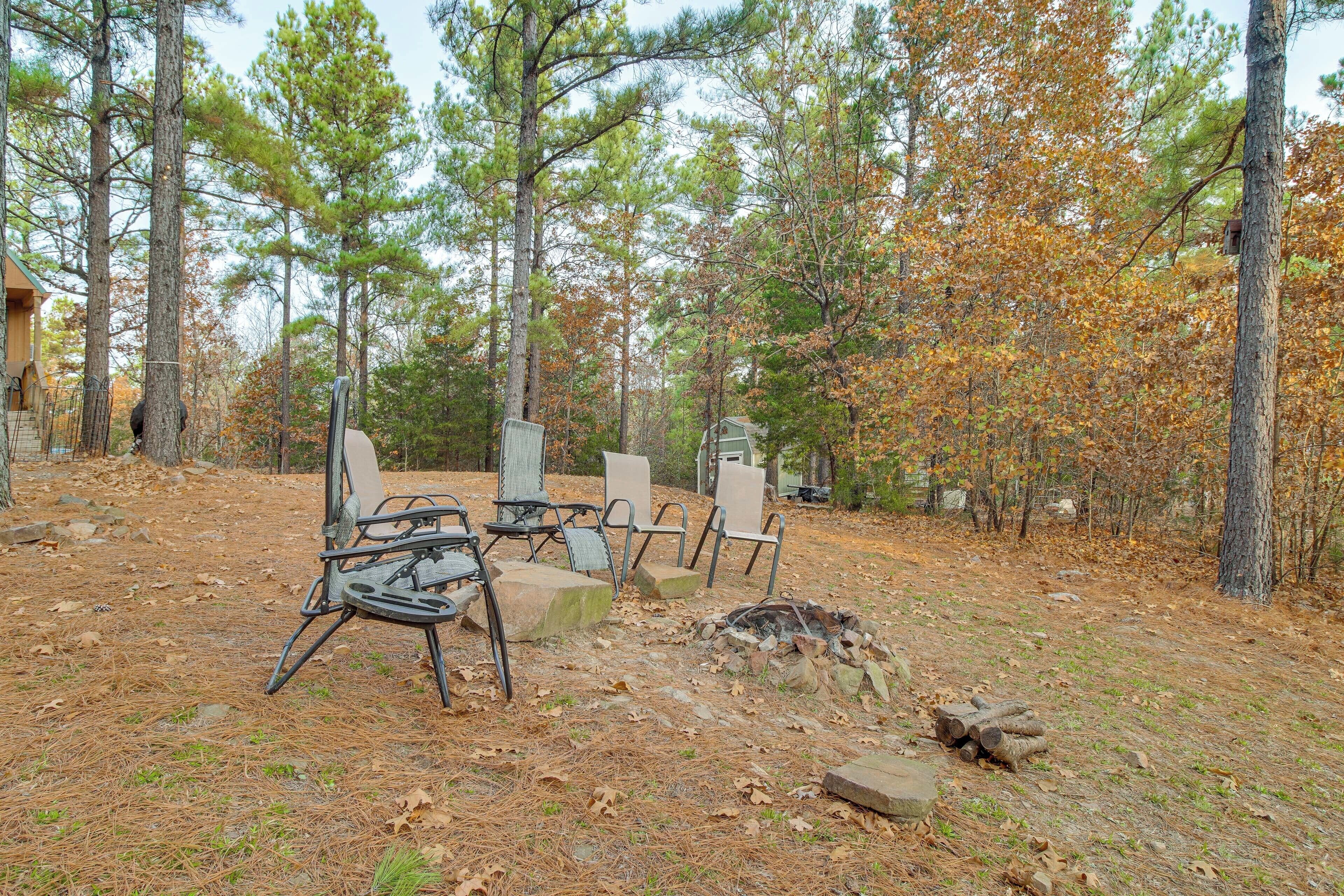Un espace extérieur aménagé dans un style rustique, avec des chaises et une table en métal, entouré d’arbres et d’une pile de bois de chauffage.
