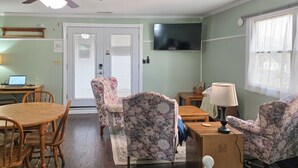 Ein Wohnzimmer mit einem Holzesstisch, floralen Ohrensesseln, einem an der Wand montierten Fernseher und einem Fenster mit Jalousien.