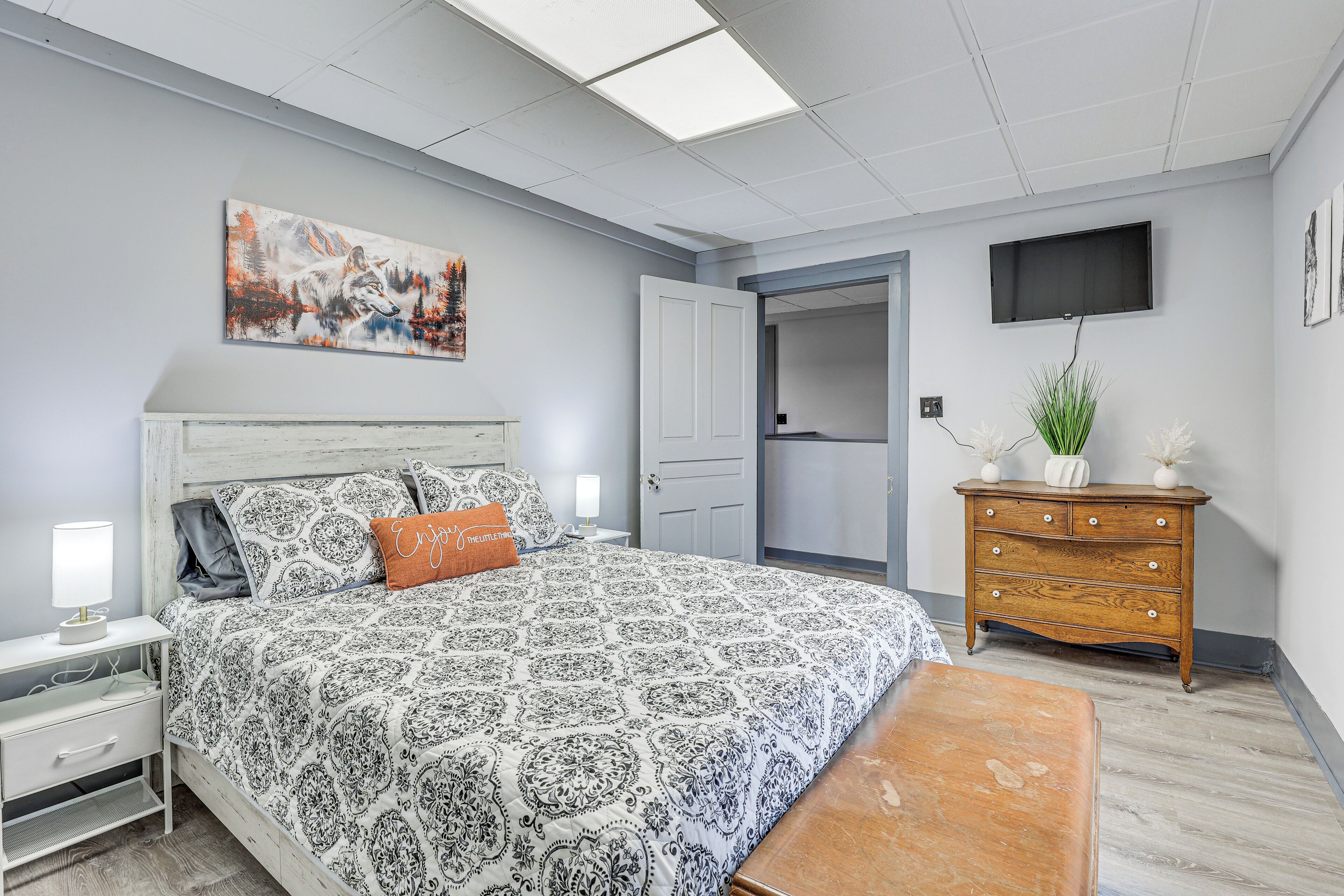 A bedroom with a patterned bedspread, a nightstand with a lamp, a dresser, a television mounted on the wall, and a door leading to another room.