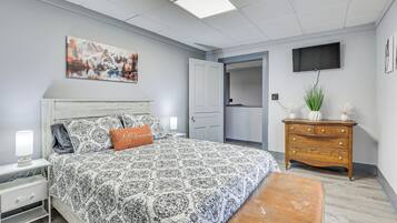 A bedroom with a patterned bedspread, a nightstand with a lamp, a dresser, a television mounted on the wall, and a door leading to another room.