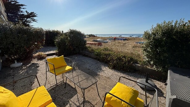 Zona salotto all'aperto con sedie gialle e un tavolo in metallo, con vista su una spiaggia e un cielo sereno, con barche.