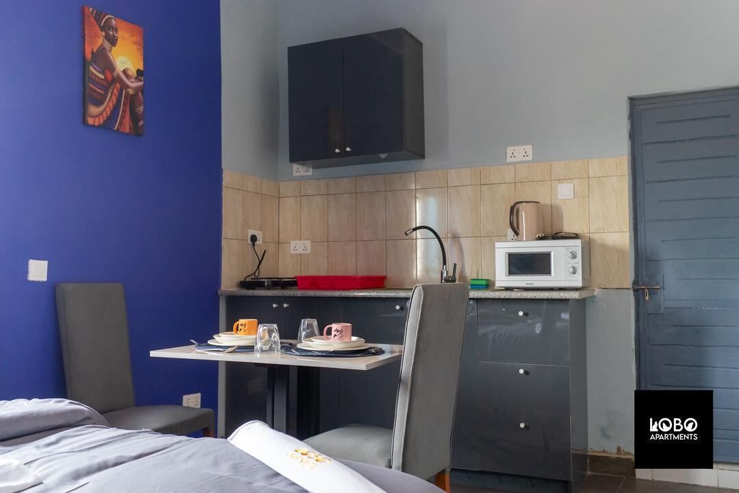 A compact room with a bed, a small dining table with chairs, a kitchenette with a microwave, and a cabinet.