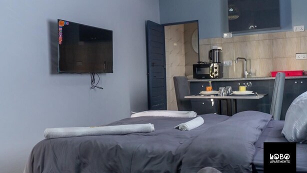 A modern bedroom with a bed, a flat-screen TV mounted on the wall, a small kitchenette, and a mirror. - Lobo studio apartments (Serrekunda)