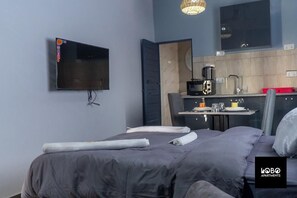 Une chambre moderne équipée d’un lit, d’un téléviseur à écran plat fixé au mur, d’une petite kitchenette et d’un miroir.