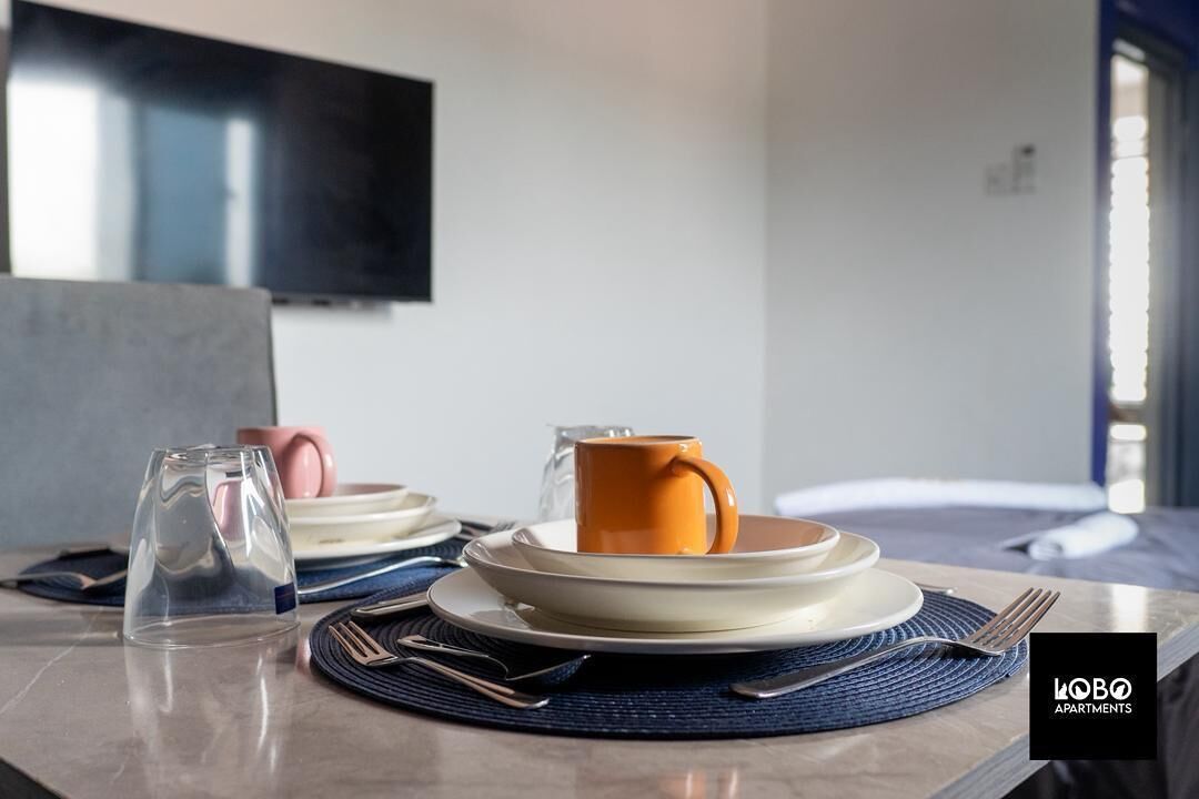 Mesa de jantar bem posta, com pratos, um copo e uma caneca, com uma televisão montada na parede ao fundo.