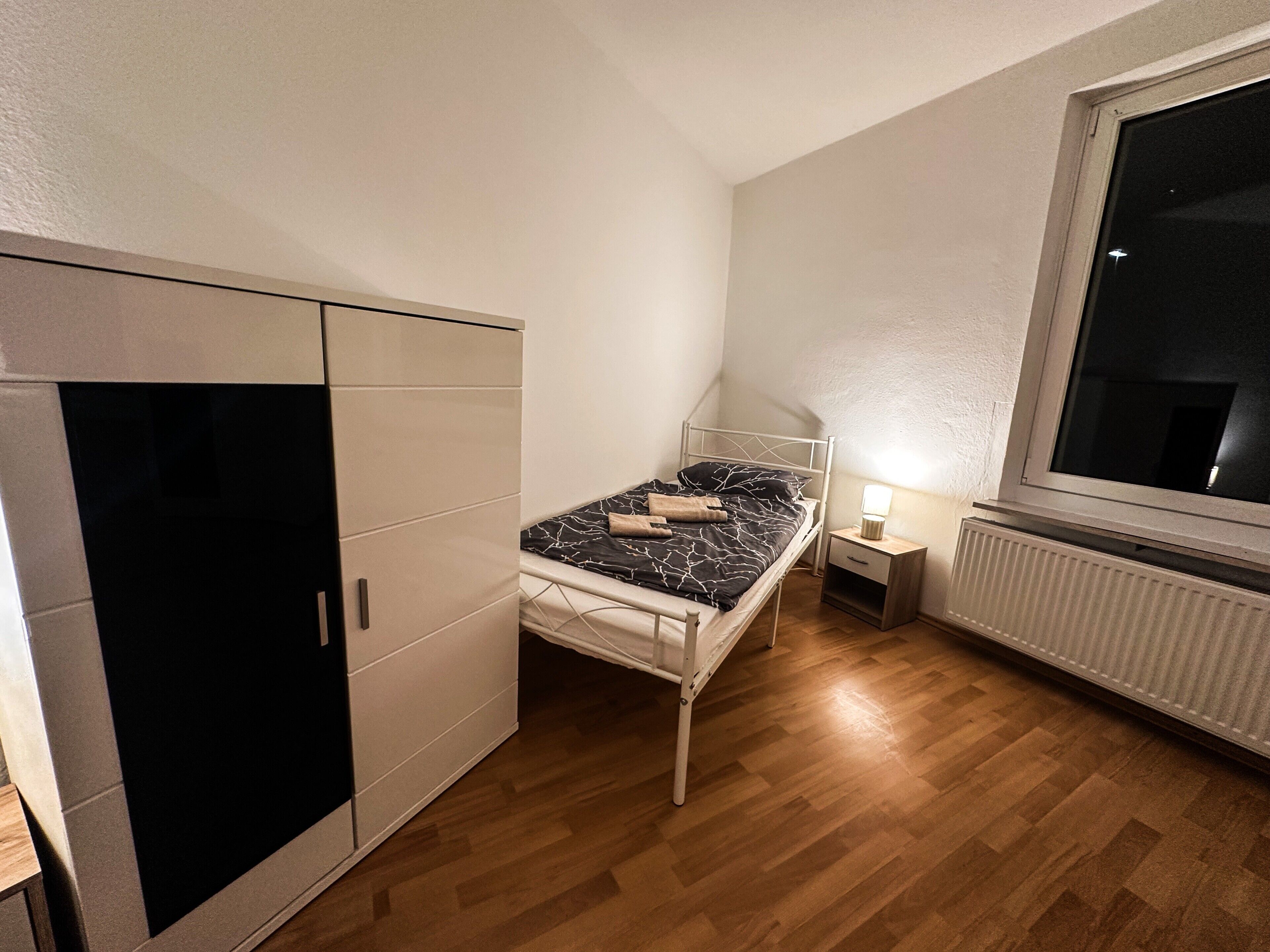 Een slaapkamer met een bed, een kledingkast, een nachtkastje, een lamp en een raam.