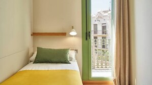Uma cama de solteiro com travesseiro verde e manta amarela, uma pequena mesa de cabeceira com abajur e vista para uma varanda com grade e edifícios ao fundo.