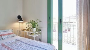 Un dormitorio con cama, mesita de noche con lámpara, planta en maceta y ventana con vista a edificios.