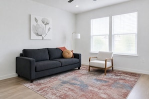 A living room with a dark grey sofa, a light-colored armchair, a patterned rug, a floor lamp, and a wall-mounted artwork. - Refershing Modern APT in A+Location|WorkerFriendly (Sherman)