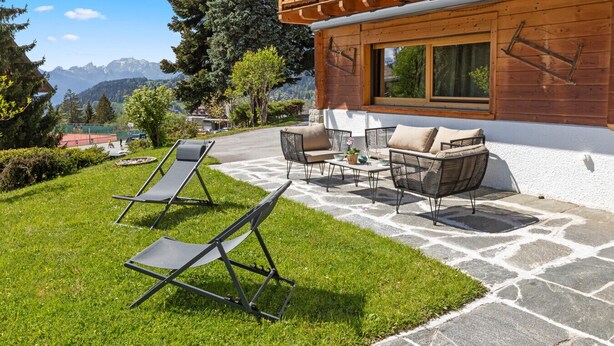 A wooden chalet with a balcony, outdoor seating, and a view of mountains.