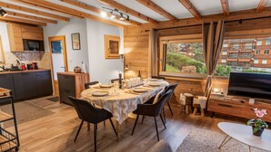 A cozy cabin-style room with a dining table set for four, a television, and a kitchen area with wooden cabinets and a window overlooking greenery.