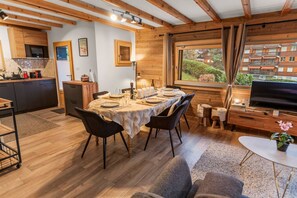 Une chambre confortable, aménagée dans le style d’un chalet, avec une table à manger pour quatre personnes, un téléviseur et un coin cuisine doté de meubles en bois et d’une fenêtre donnant sur un espace verdoyant.