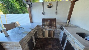 Una cocina al aire libre con una parrilla empotrada, armarios de acero inoxidable y suelo de baldosas de piedra.