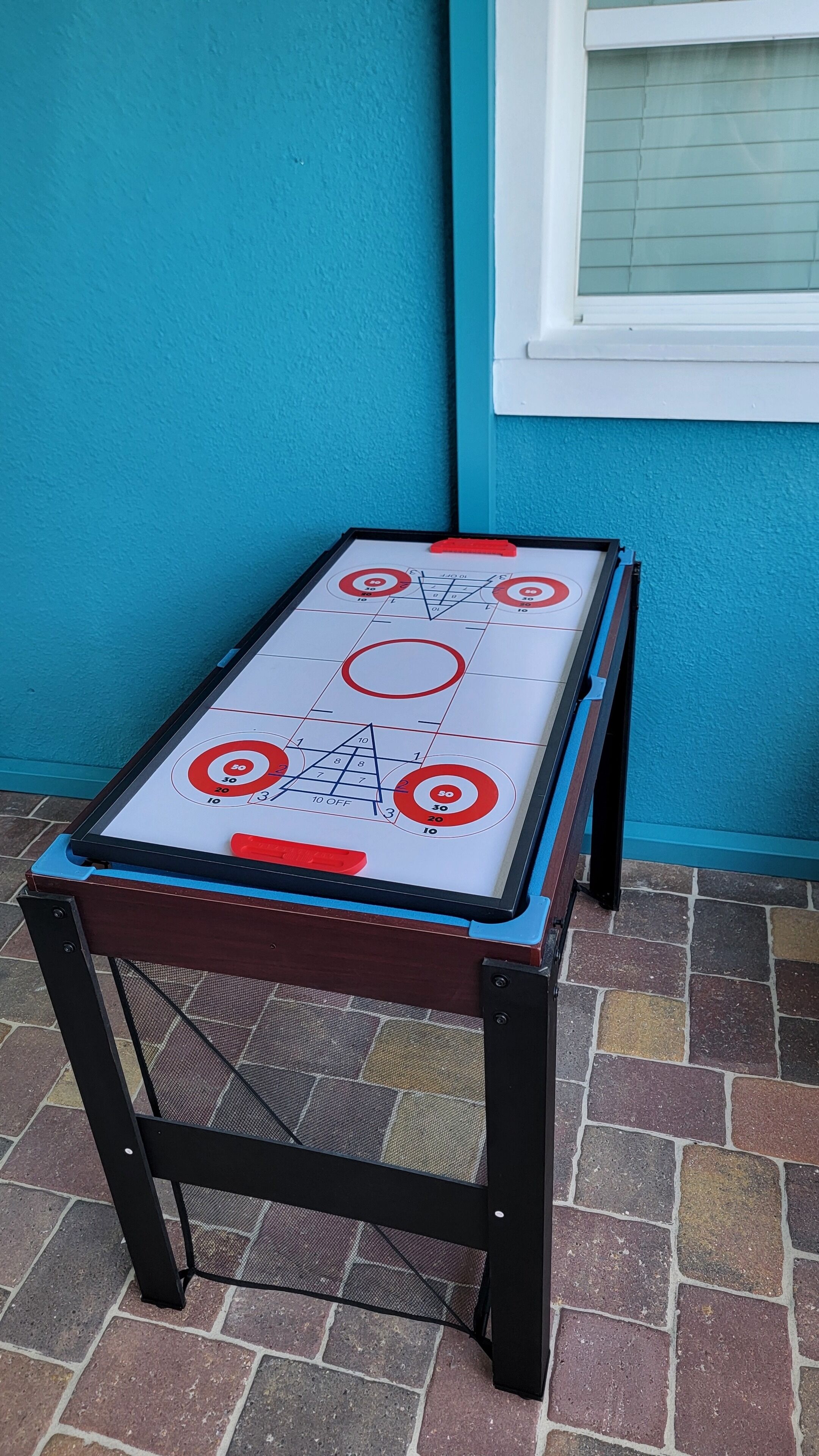 A shuffleboard table with a target and numbered scoring areas, placed on a brick patio against a blue wall.