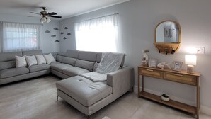 Una sala de estar moderna con un sofá seccional gris, un pequeño otoman, una mesa consola de madera con objetos decorativos y un ventilador de techo.