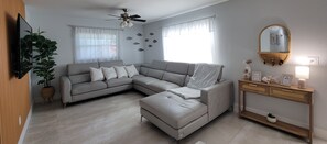 A modern living room with a grey sectional sofa, a small ottoman, a wooden console table with decorative items, and a ceiling fan. - Villa Azalea · 4BR Heated Pool · Game Room · Near Gulf Beaches (Seminole)