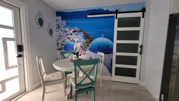 Una habitación con vistas al mar azul y edificios blancos, una mesa con sillas y una puerta corredera.